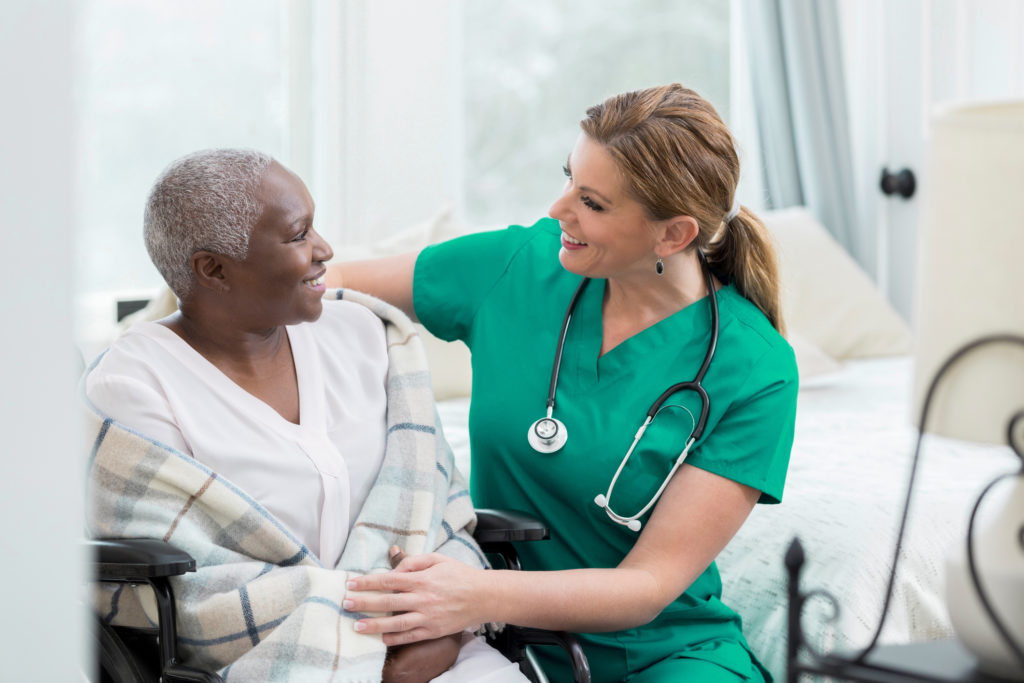 Mature Caucasian female nurse assists a female patient in the patient's home. The patient is in a wheelchair.